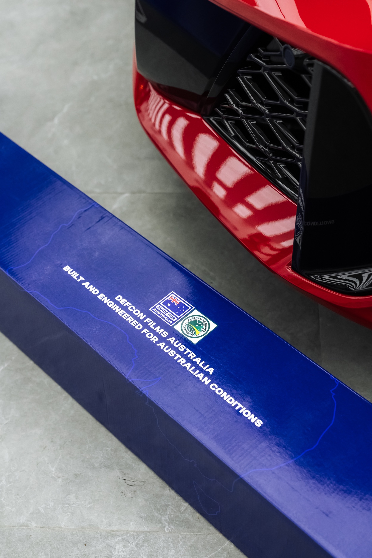 Front bumper of a red performance car beside a Defcon Films Australia paint protection film box marked made for Australia