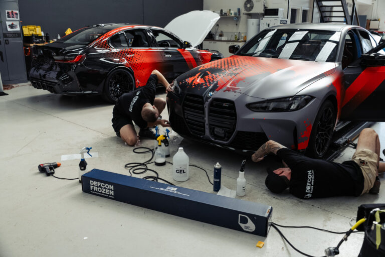 Two installers wrapping BMW M cars with red and black livery in a workshop with a Defcon Films Australia Defcon Frozen satin paint protection film box